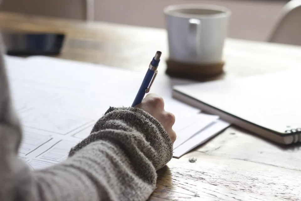 Person writing notes on paper beside a cup of coffee