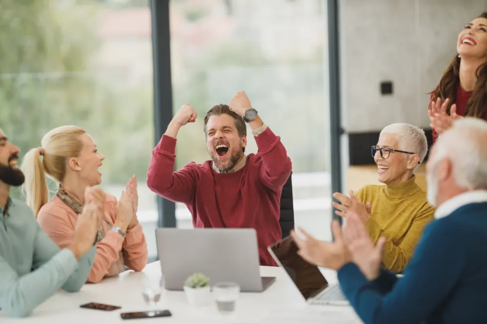 Colleagues cheering around a laptop in a bright meeting room