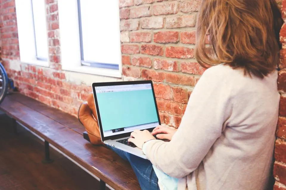 Woman working on a laptop beside a brick wall and window